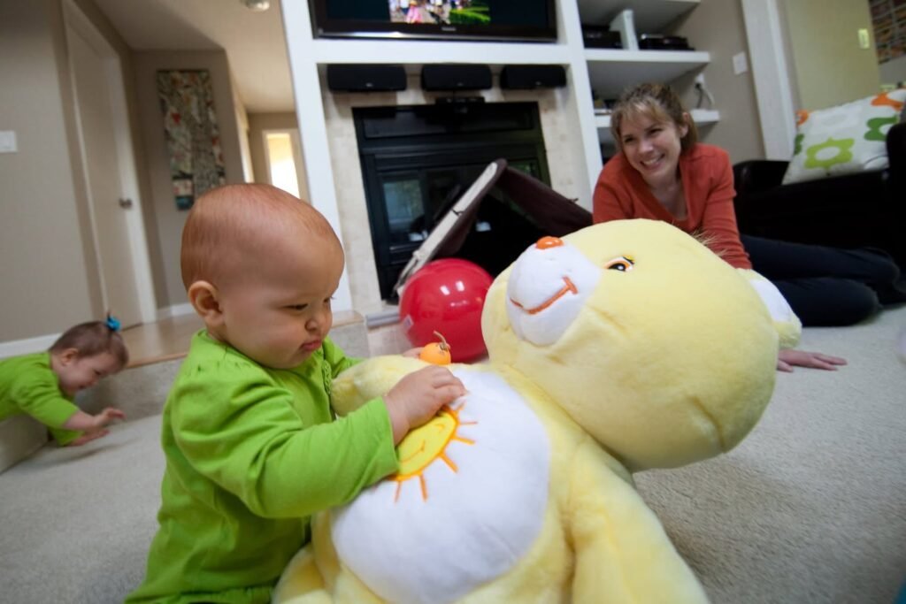 A young toddler in a green long-sleeve shirt interacts with a giant yellow teddy bear in a warm living room. An adult sits nearby on the floor smiling, capturing a candid family moment.