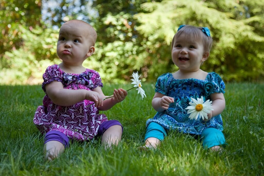Two toddlers sitting on a lawn in matching blue and purple outfits, holding white daisy flowers.