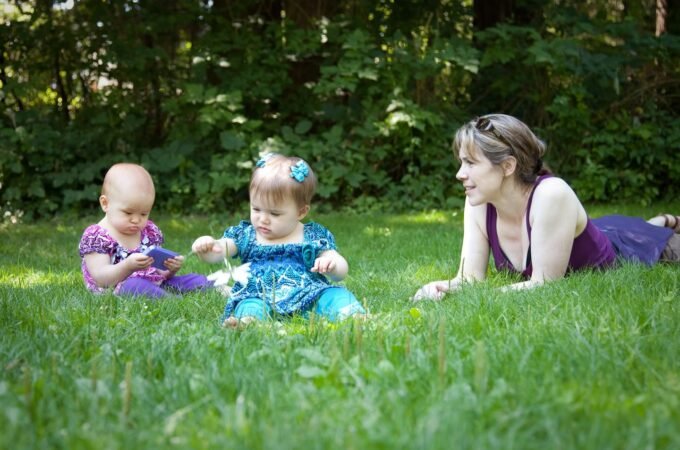 A woman and two young children enjoying a sunny day on the grass, surrounded by lush green trees.