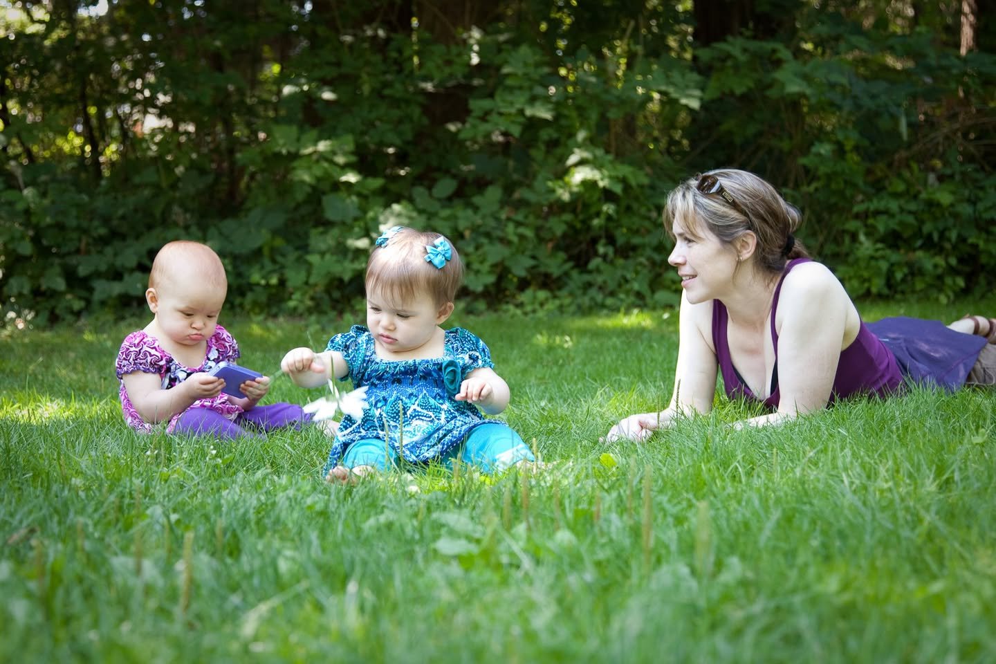 11 Months A woman and two young children enjoying a sunny day on the grass, surrounded by lush green trees.
