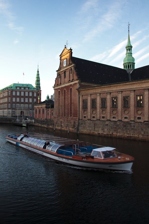 A vintage boat glides along a calm waterway beside ornate red-brick buildings and green copper spires.