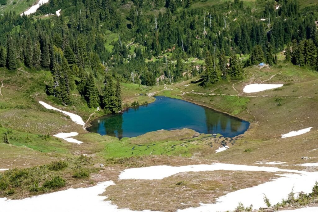 A serene view of a mountain lake surrounded by dense evergreen forests and lingering patches of snow.