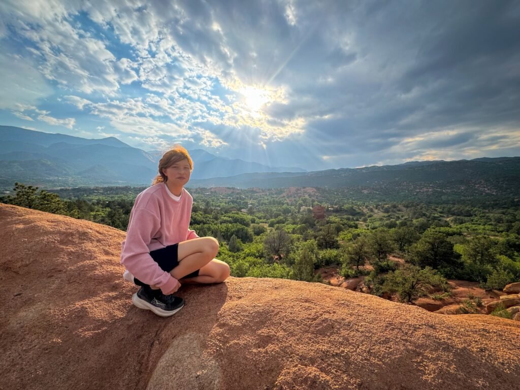 Kristie sits on a rocky ledge overlooking a stunning mountainous landscape with a vibrant sunset sky. She is wearing a pink top and dark shorts, soaking in the serene moment.