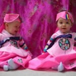 High Divide Loop Two young children wearing traditional pink hanbok with colorful accents. The outfit features a bright pink dress matching a vibrant curtain backdrop.