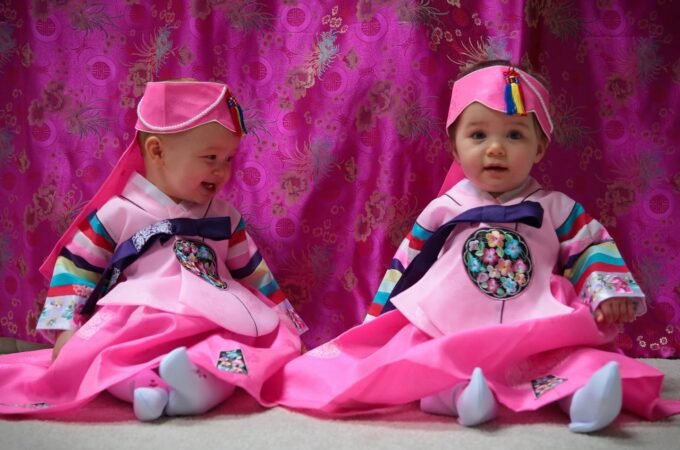 Two young children wearing traditional pink hanbok with colorful accents. The outfit features a bright pink dress matching a vibrant curtain backdrop.