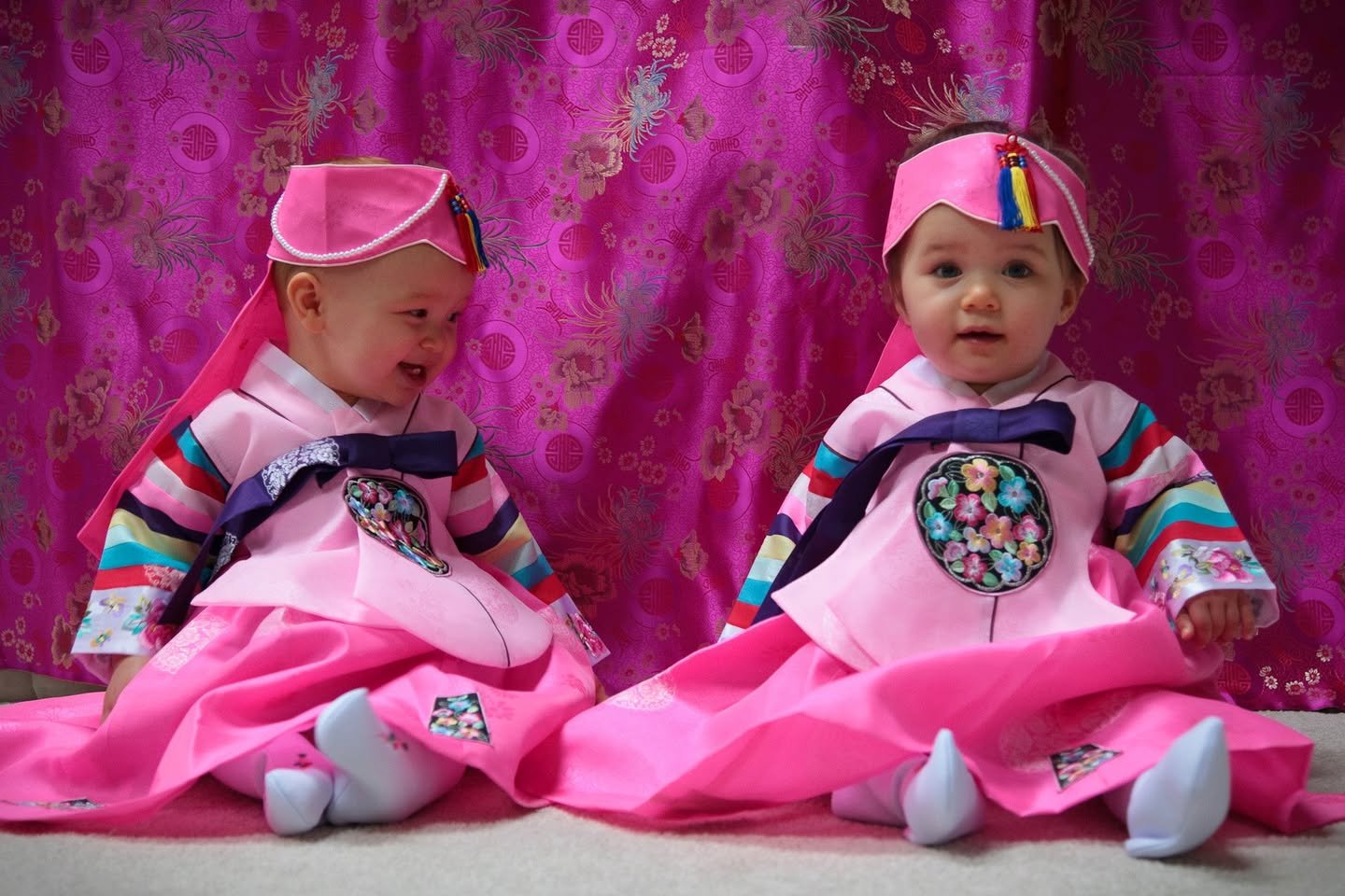 Two young children wearing traditional pink hanbok with colorful accents. The outfit features a bright pink dress matching a vibrant curtain backdrop.
