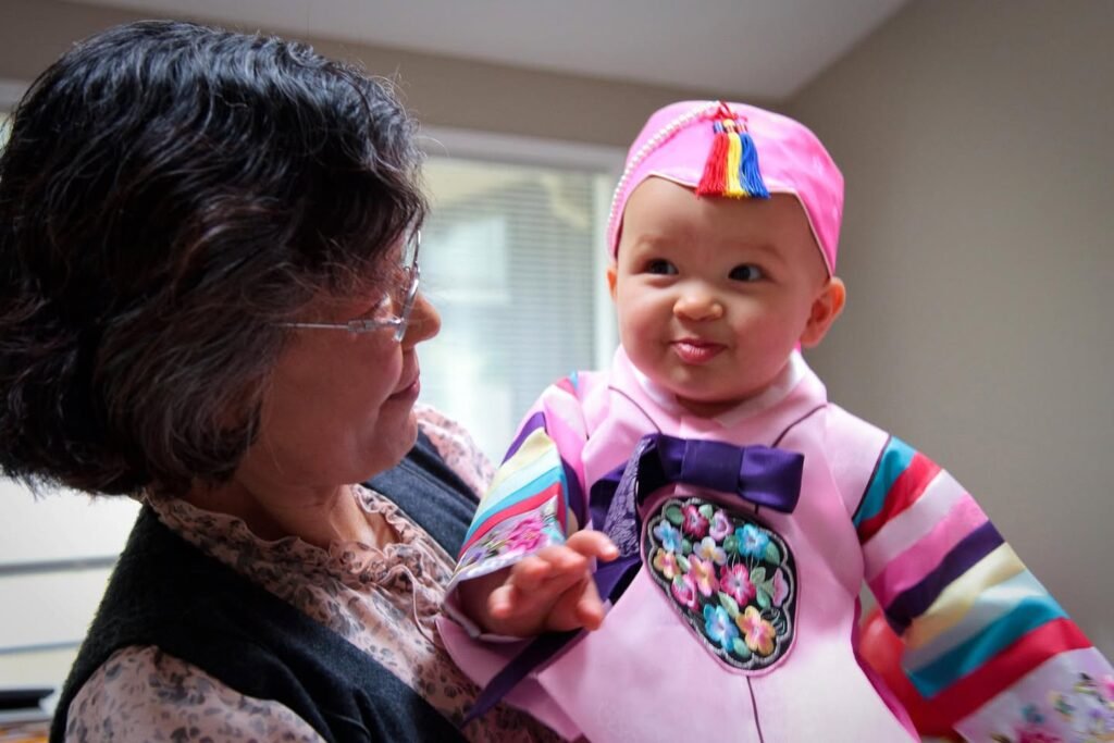 A grandmother holds her granddaughter who is dressed in a colorful striped outfit and matching cap. The baby smiles while the older woman looks on affectionately in an indoor setting.
