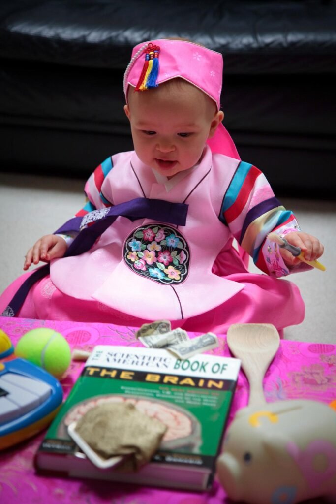 A toddler in a colorful costume sits in a play seat with toys and a book on a pink blanket.