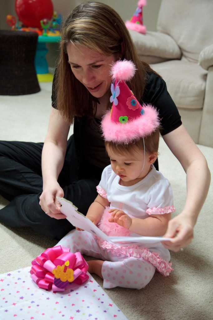 A mother helps a toddler put on a pink party hat. The young girl looks down at her feet.