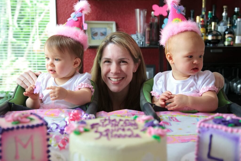 A woman and two young children wearing pink party hats sit together at a table with decorative birthday cakes. The scene is indoors with floral tablecloths and colorful decorations.