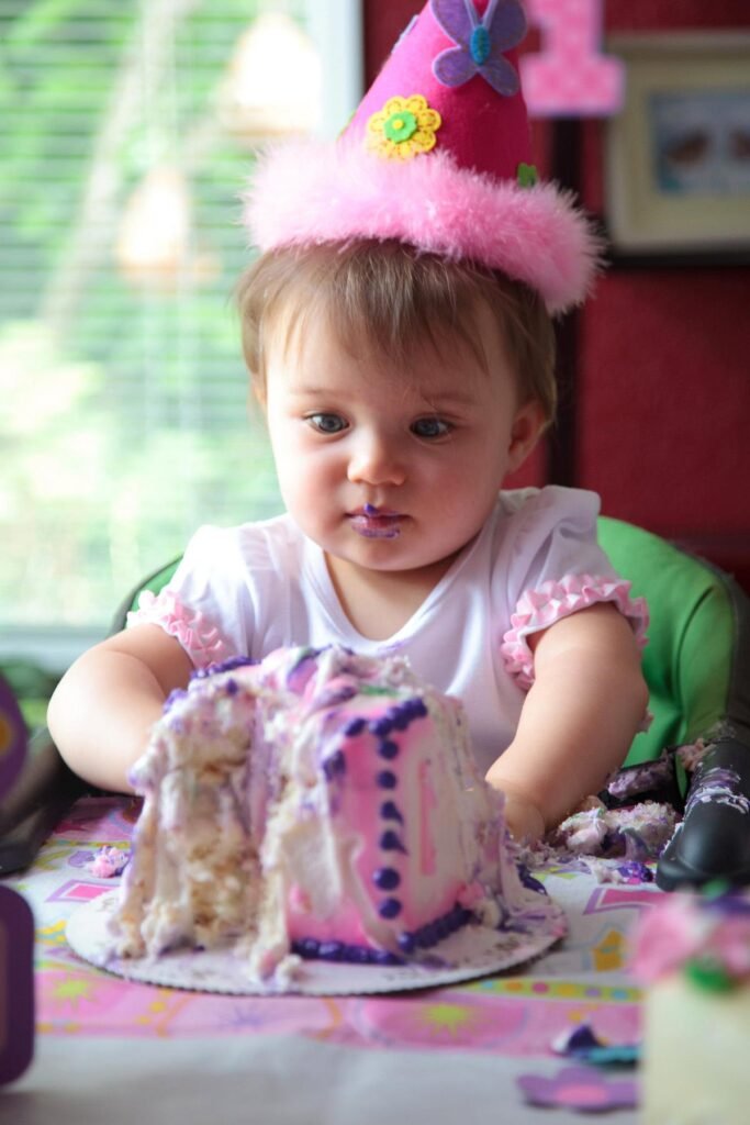 A child wearing a pink number one party hat sits in a green highchair with a pink and purple birthday cake.