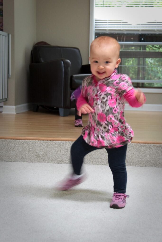 A toddler in a floral pink shirt happily dances on a gray carpet. The motion blur captures the energy and joy of the moment indoors.