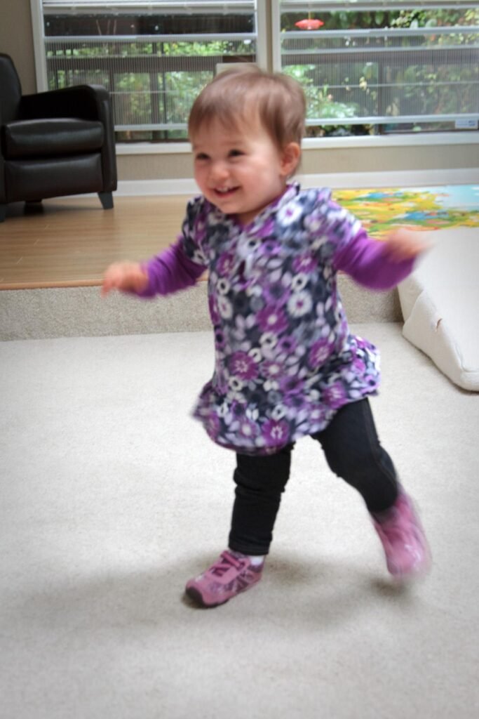 A toddler in a floral tunic dances indoors with arms outstretched. A close-up captures the joyful, blurred movement.