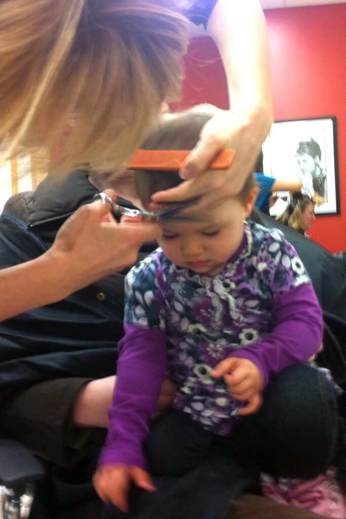A young child in a purple shirt gets a haircut in a salon with red walls. The scene captures a quiet, intimate moment of personal care.