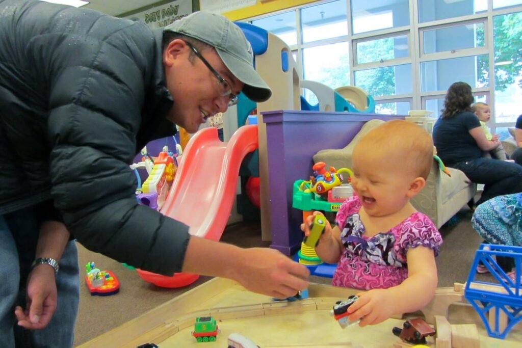 An adult wears a black puffer jacket and gray cap, gently interacting with a toddler in a pink dress at a wooden play table.