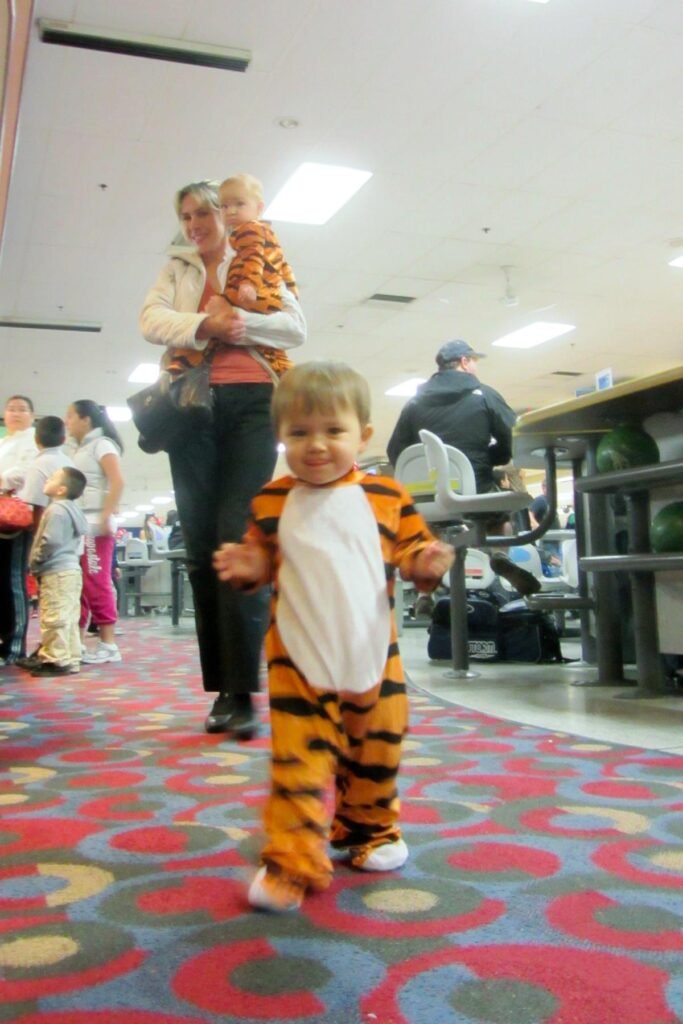 A toddler in a tiger-themed costume walking through a busy public indoor space with colorful patterned carpet.