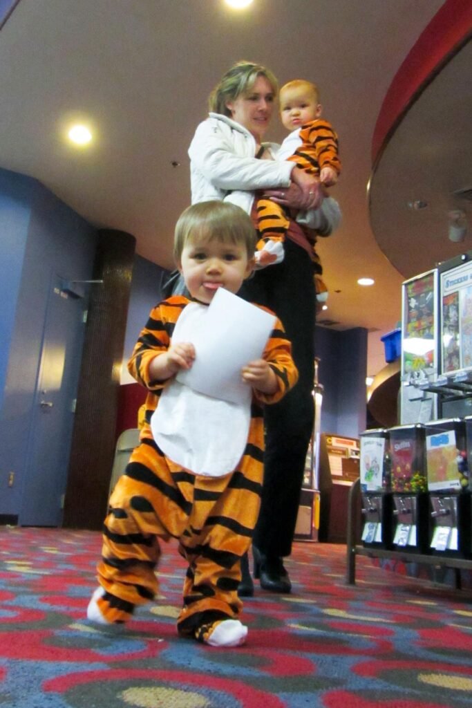 Small child in a tiger pajama costume holding a piece of paper in a movie theater lobby with an adult standing nearby.