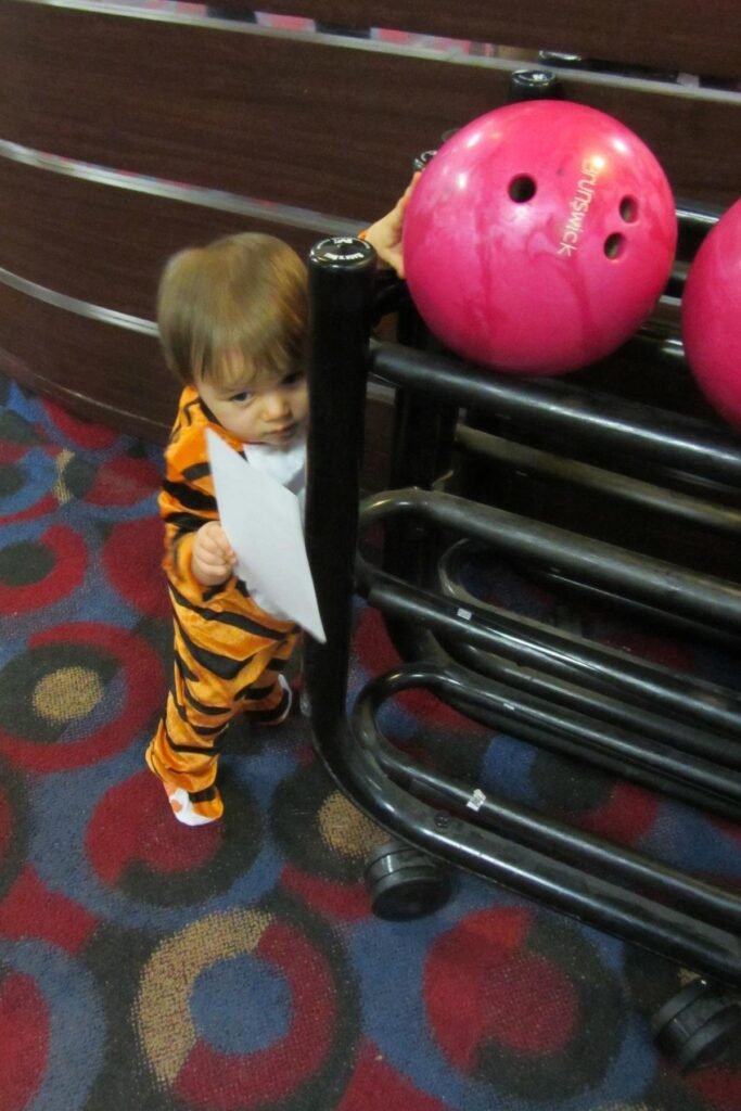 A toddler in a tiger costume stands near a pink bowling ball on the ball return rack.