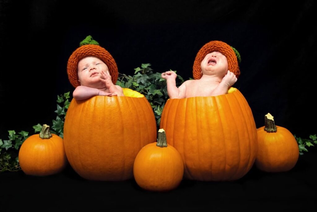 Two children wearing pumpkin-themed hats sit inside large pumpkins surrounded by a festive autumn arrangement.