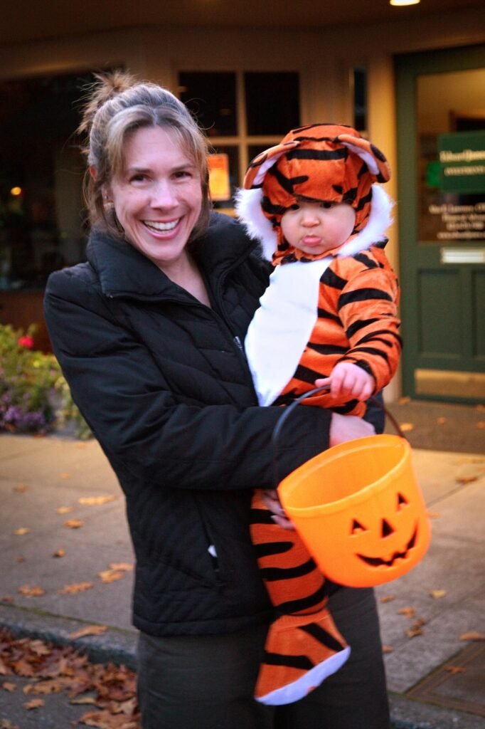 A smiling adult holds a child dressed in a full tiger costume with an orange pumpkin bucket. They stand on a front porch with autumn leaves on the ground.