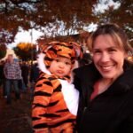 Safari: Part 2 of 3 A woman smiles while holding a child in a tiger costume at an outdoor Halloween event. The background shows autumn trees and a festive crowd.