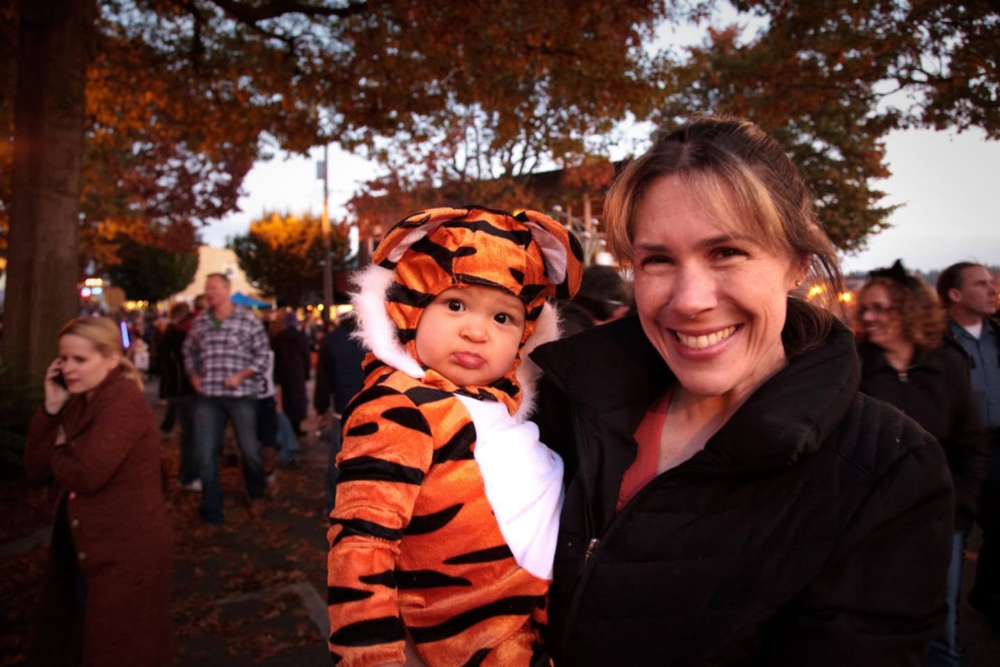 A woman smiles while holding a child in a tiger costume at an outdoor Halloween event. The background shows autumn trees and a festive crowd.