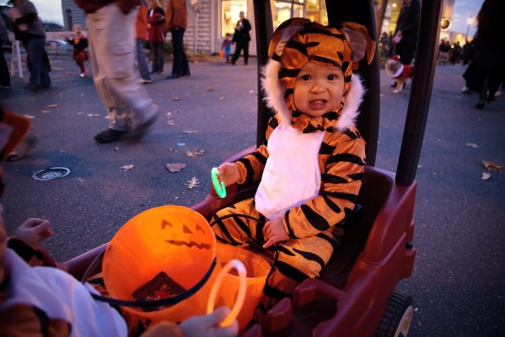 A child dressed in a tiger costume sits in a wagon during a Halloween parade while holding a carved pumpkin and a glow bracelet.