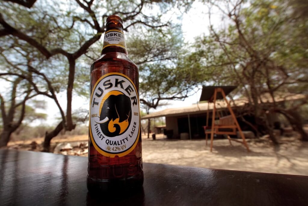 A cold Tusker beer bottle sits on a dark table in the foreground, with blurred safari trees and a wooden lodge in the background.