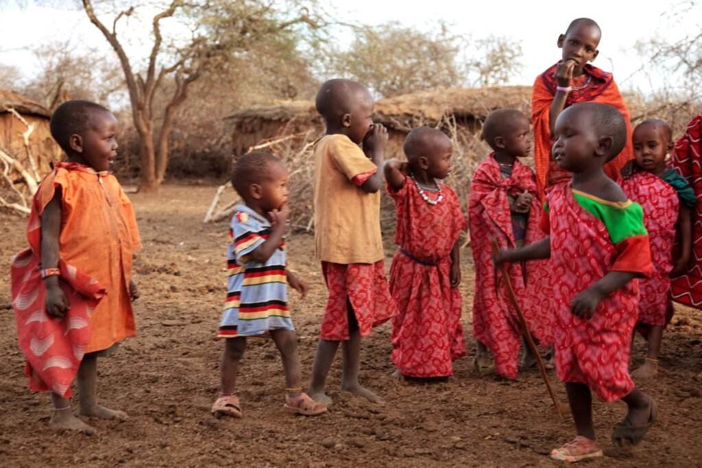 A group of children in colorful traditional clothing stand in a rural village setting with dirt ground and a thatched hut in the background.