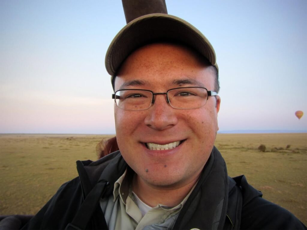 A selfie taken from a hot air balloon basket overlooking a vast grassland with another balloon in the distance.