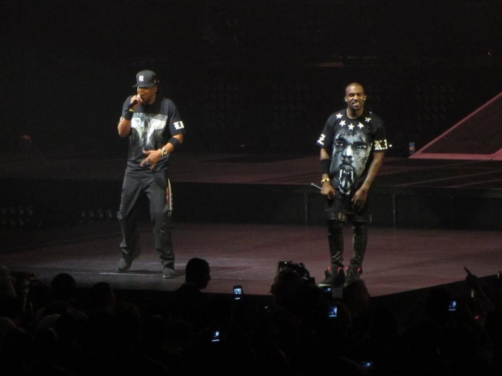 Two performers on a concert stage with an engaged audience in the foreground. The stage is dimly lit with moody stage lighting.