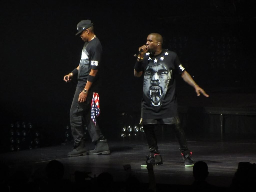Kanye West performs on stage wearing a black t-shirt with a face graphic, singing into a microphone during a live concert.
