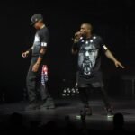 Kanye West performs on stage wearing a black t-shirt with a face graphic, singing into a microphone during a live concert.