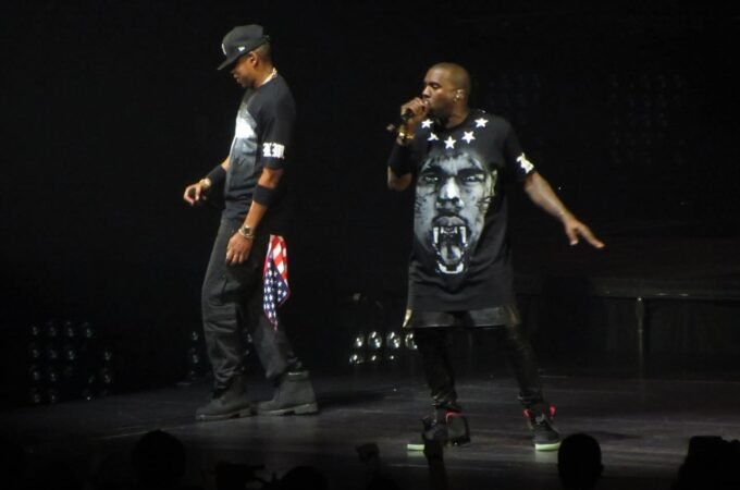 Kanye West performs on stage wearing a black t-shirt with a face graphic, singing into a microphone during a live concert.