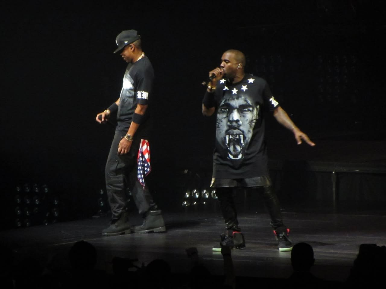 Kanye West performs on stage wearing a black t-shirt with a face graphic, singing into a microphone during a live concert.