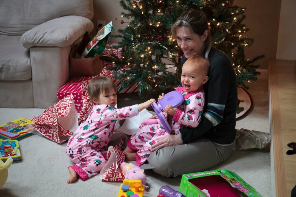 A woman and young children in matching pajamas gather around a Christmas tree with presents on the floor. The image captures a cozy holiday moment in a living room.