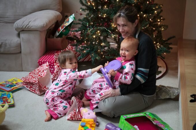 A woman and young children in matching pajamas gather around a Christmas tree with presents on the floor. The image captures a cozy holiday moment in a living room.