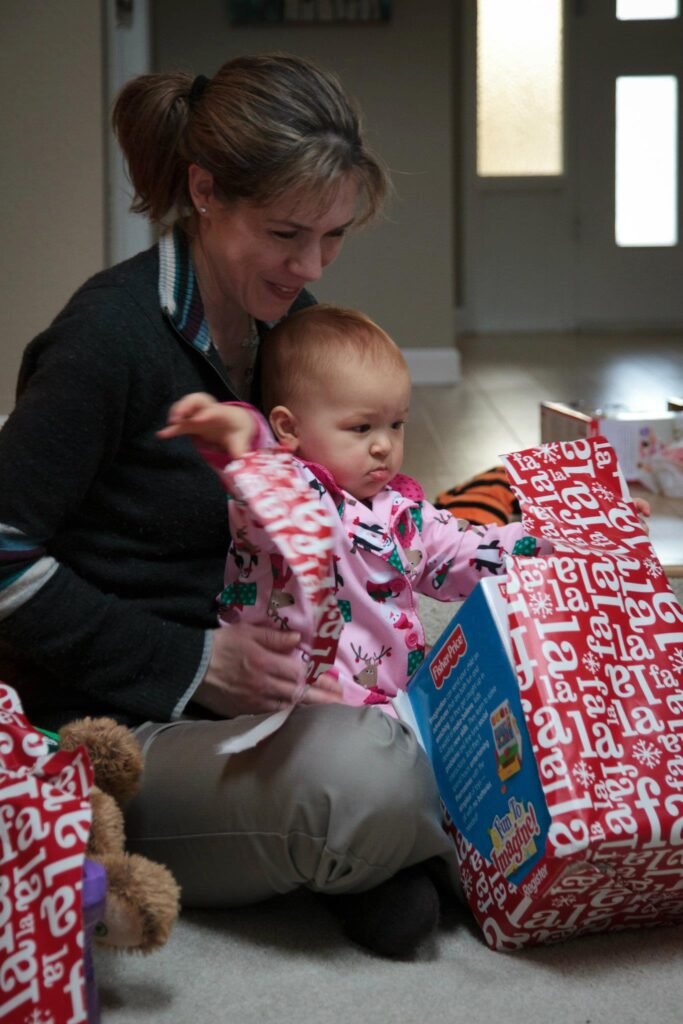 A baby opens a red Christmas gift bag while an adult smiles nearby.