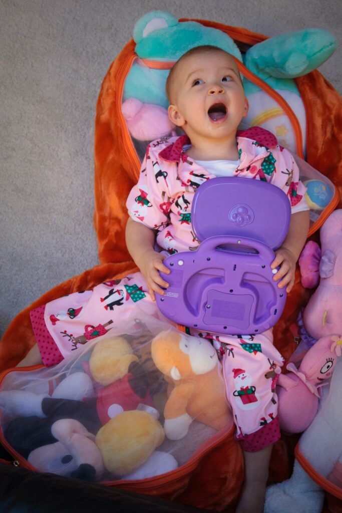 A happy baby in holiday pajamas holds a purple toy activity center while sitting in an orange and teal padded bouncer surrounded by plush stuffed animals.
