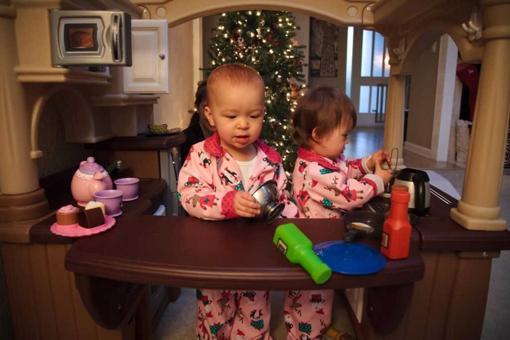 Two young children in matching pajamas playing with toy food in a wooden play kitchen with a Christmas tree in the background.