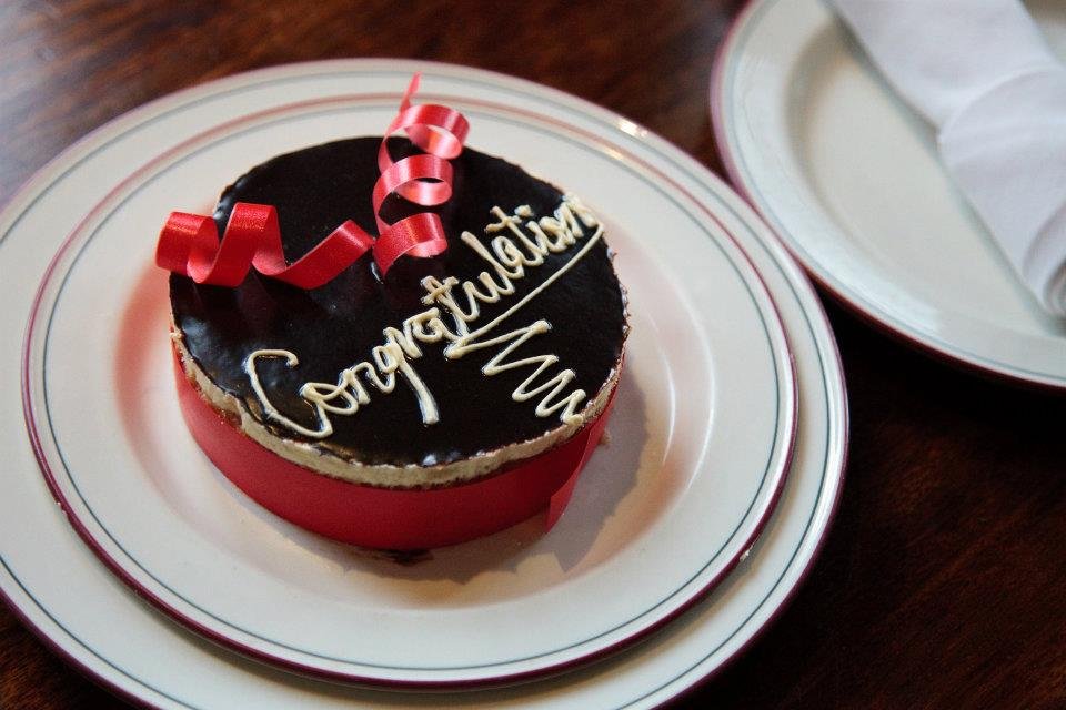 A close-up of a small chocolate cake with a red ribbon and 'Congratulations' written in white.