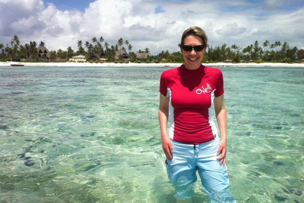 A person standing in shallow, crystal-clear water with a tropical island and palm trees in the background.