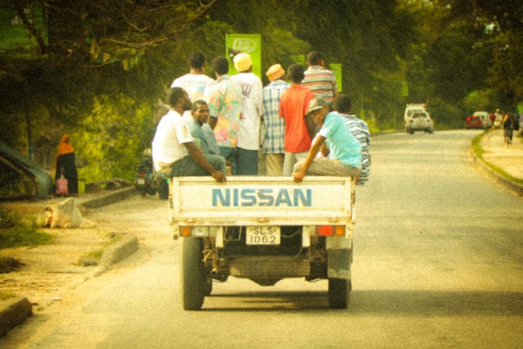 People riding in the back of a white Nissan pickup on a dusty road.