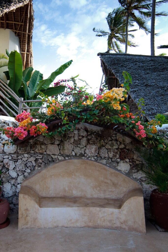 A tropical garden with vibrant flowers and lush greenery surrounding a rustic stone building. Palm trees sway against a bright blue sky, creating a serene atmosphere.