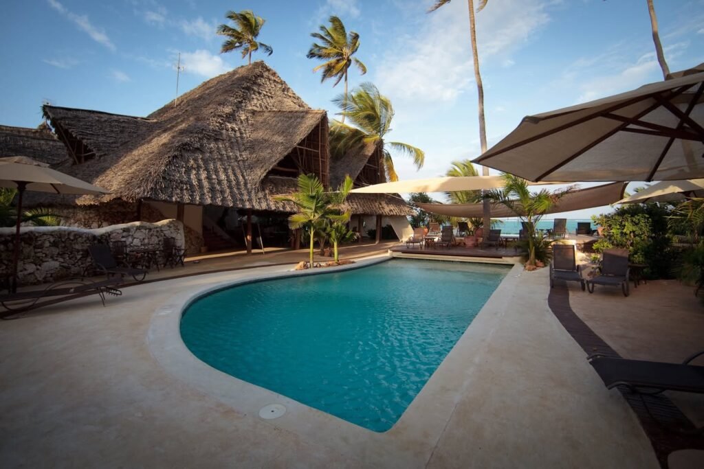 Serene tropical resort featuring a crystal-clear swimming pool, thatched-roof villas, and lush palm trees under a bright blue sky.