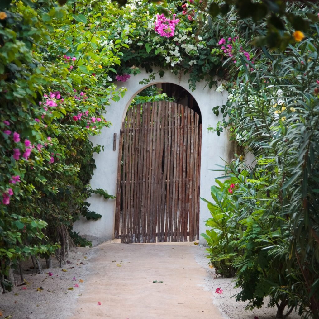 A compact arched gate made of vertical reeds framed by a white wall, with vibrant green foliage and bright pink flowers growing on either side.