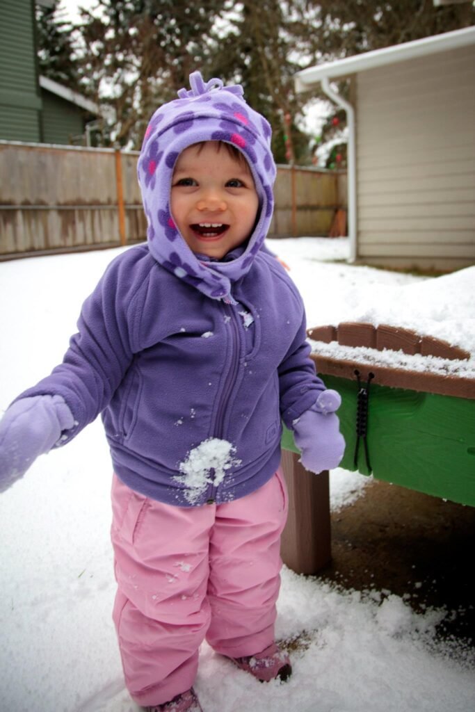 A small child in a purple hat and pink pants stands in the snow, smiling joyfully.