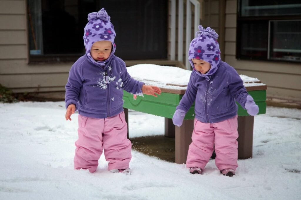Two toddlers in matching purple hats and pink snowsuits stand in the snow next to a green table.