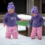 Two toddlers in matching purple hats and pink snowsuits stand in the snow next to a green table.