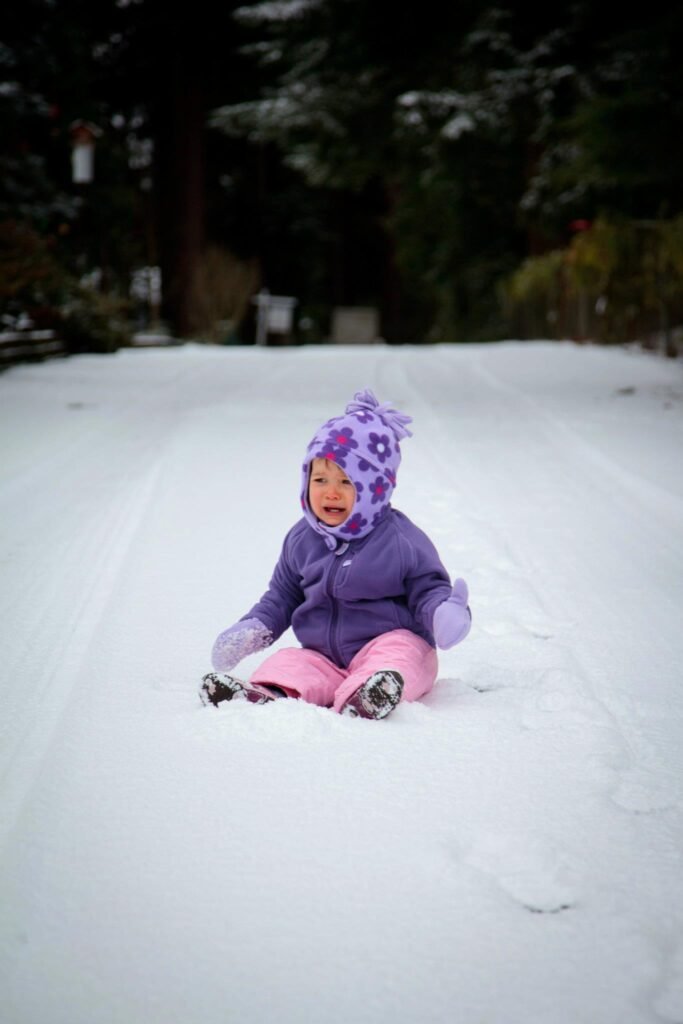 A child bundled in warm winter clothes happily slides down a snowy hill in the Pacific Northwest surrounded by snow-covered trees.